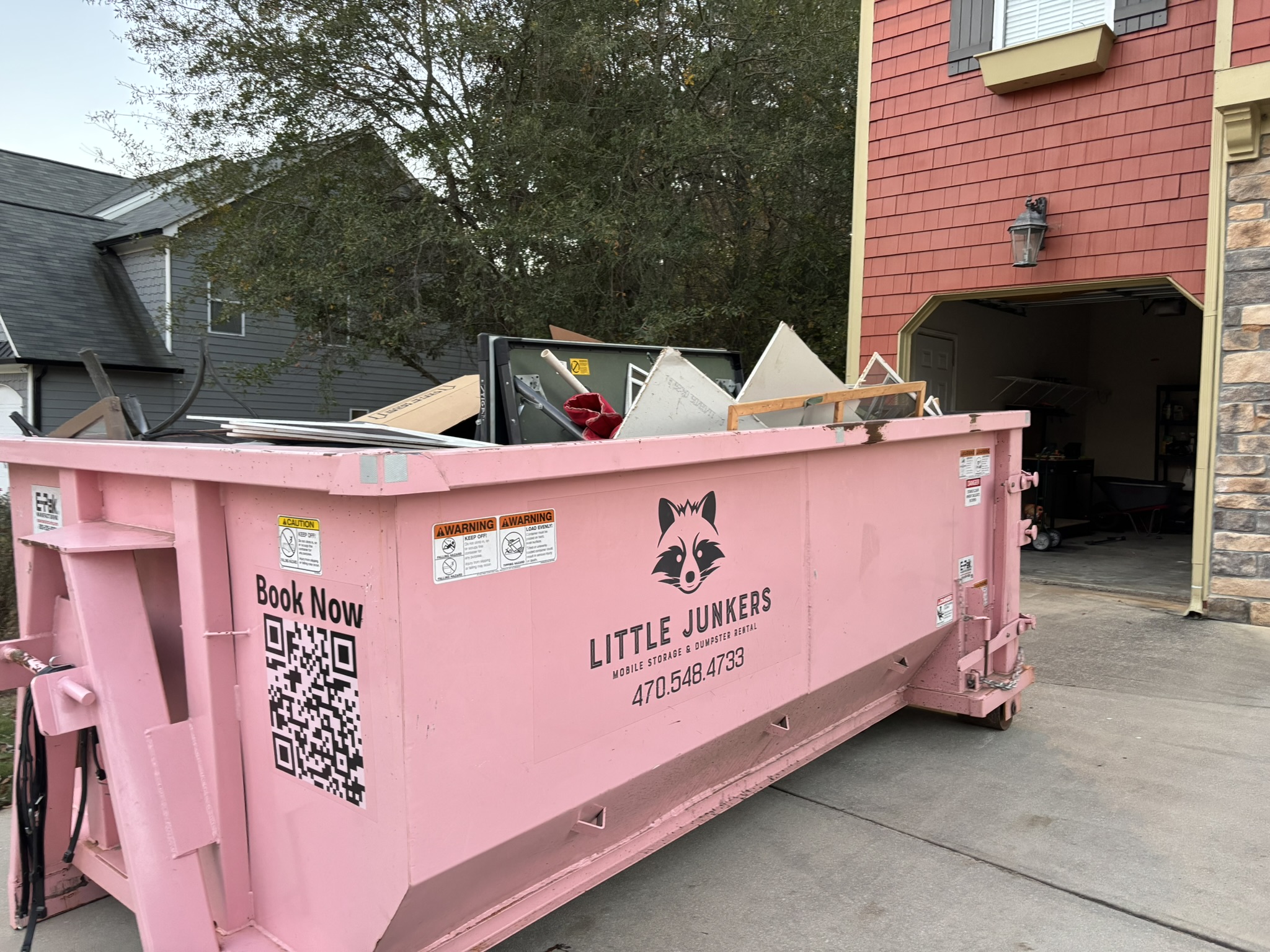 Pink 11-yard Little Junker dumpster loaded beside a residential garage